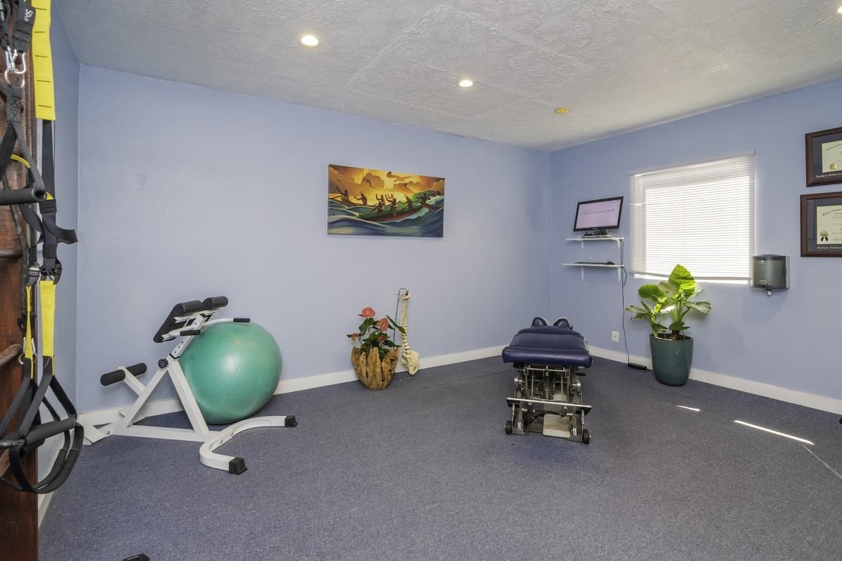 A bright physical therapy and rehab room at Kailua Wellness Center with treatment tables, an exercise ball and a plant